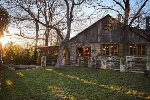 A rustic stone cabin surrounded by trees, with sunlight streaming through, casting long shadows on the grassy lawn.