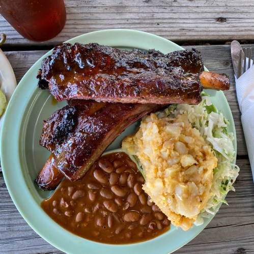 A plate with glazed ribs, baked beans, and creamy potato salad on a wooden table.