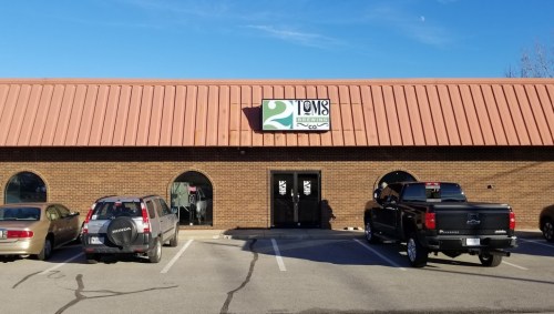 Exterior of a brick building with a sign reading "2 Toms Brewing" and parked cars in front. Clear blue sky above.