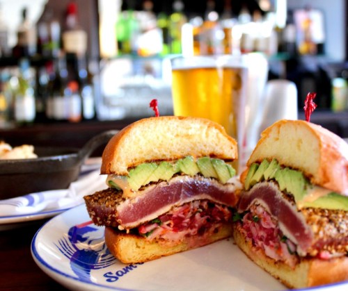 A close-up of a sandwich filled with seared tuna, avocado, and pink sauce, served on a plate with a beer in the background.