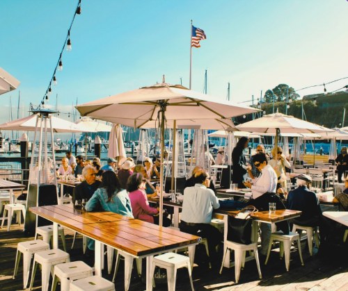A bustling outdoor restaurant by the marina, with diners under umbrellas and sailboats in the background.