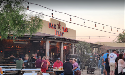 A lively outdoor scene at San Van Flat, with people socializing and string lights overhead during sunset.