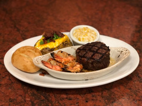 A plate featuring a grilled steak, shrimp, a loaded baked potato, corn, and a roll on a marble table.