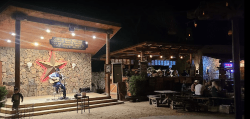 A musician performs on stage at a rustic outdoor venue with a bar and seating, illuminated by warm lights at night.