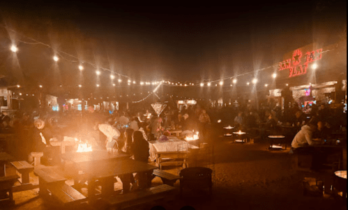 A lively outdoor gathering at night, with string lights and fire pits, filled with people enjoying the atmosphere.