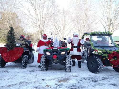 A festive group in winter attire poses with decorated ATVs, celebrating the holiday season in a snowy landscape.