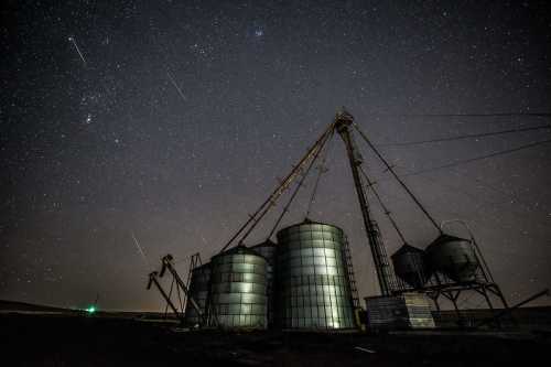 A silhouette of grain silos under a starry night sky, with shooting stars visible in the background.