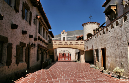 A sunlit courtyard featuring rustic buildings with arched doorways and a clock tower in the background.