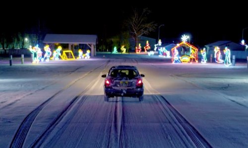 A car drives through a snowy area, passing colorful holiday lights and decorations at night.