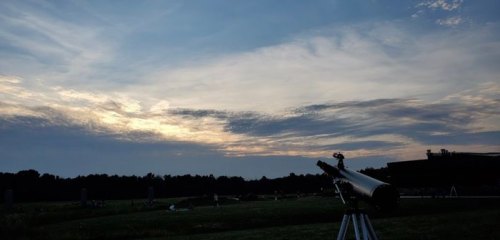 A telescope on a tripod facing a cloudy sky at dusk, with a grassy field and trees in the background.