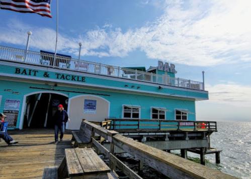 A wooden pier extends towards the water, with a turquoise building labeled "Bait & Tackle" and an American flag.
