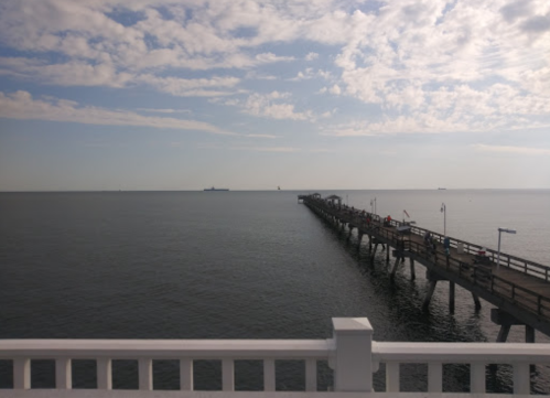 A wooden pier extends into calm waters under a partly cloudy sky, with distant ships visible on the horizon.