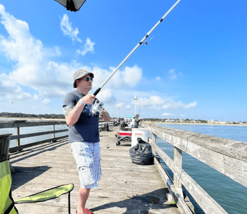 A person fishing from a pier on a sunny day, wearing a hat and shorts, with boats and a clear blue sky in the background.