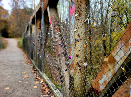A weathered bridge with a chain-link fence adorned with colorful love locks, surrounded by autumn foliage.