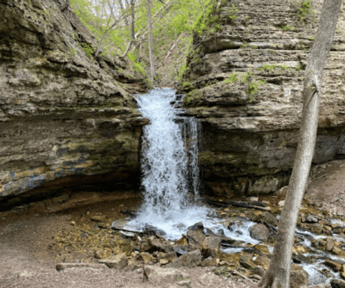 A small waterfall cascades over rocky cliffs, surrounded by lush greenery and trees in a serene natural setting.