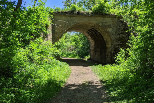 A stone archway surrounded by lush greenery, leading to a dirt path through a vibrant, wooded area.