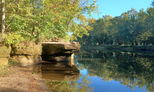 A serene river scene with rocky banks, surrounded by trees reflecting in the calm water under a clear blue sky.