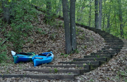 Two blue kayaks are resting on a leaf-covered path beside wooden steps in a lush green forest.