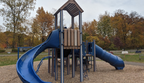 A blue playground structure with slides and climbing features, surrounded by autumn trees and a gravel surface.