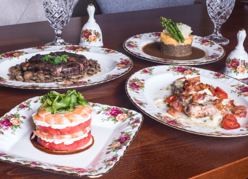 A beautifully arranged table with four gourmet dishes on floral plates, featuring steak, seafood, and a layered dessert.