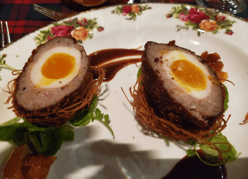 Two scotch eggs on a decorative plate, with a golden yolk center, surrounded by greens and a drizzle of sauce.