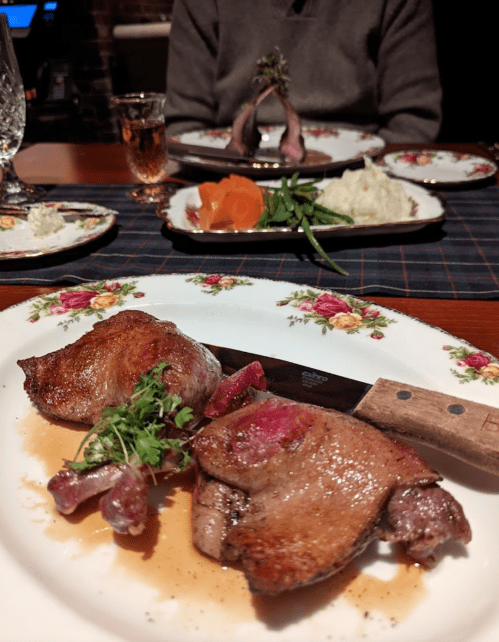 A plate with two pieces of roasted meat garnished with herbs, a knife, and a table set with additional dishes in the background.