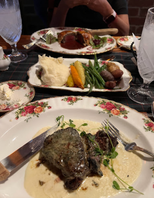 A beautifully plated meal featuring steak, mashed potatoes, vegetables, and garnishes, with drinks in the background.