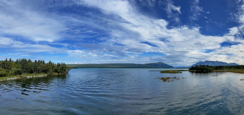 A panoramic view of a serene lake surrounded by lush greenery and distant mountains under a blue sky with clouds.