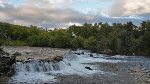 A serene river with a waterfall, surrounded by lush greenery and a cloudy sky in the background.