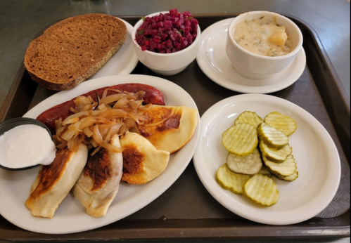 A tray with sausage, pierogi topped with onions, pickles, beet salad, rye bread, and a bowl of creamy soup.