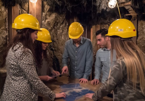 A group of five people in yellow hard hats examines a map on a table in a dimly lit, cave-like setting.