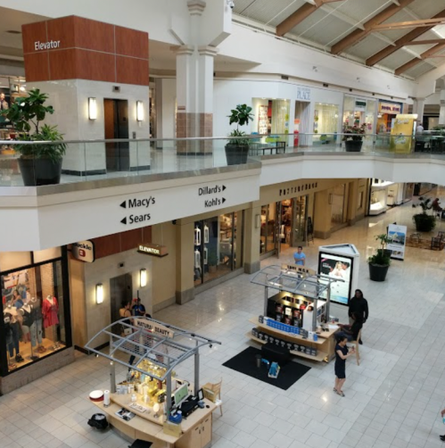 A spacious mall interior with shops like Macy's and Dillard's, featuring a central kiosk and shoppers.