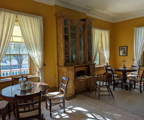 A cozy room with yellow walls, wooden furniture, and large windows letting in natural light.