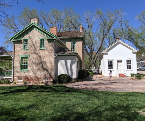 A historic two-story house with green trim next to a smaller white building, surrounded by trees and grass.