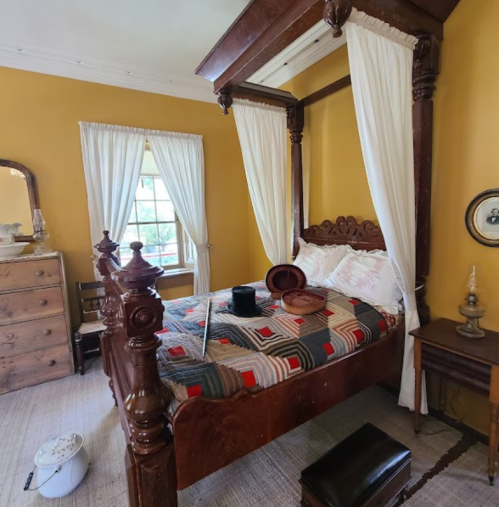 A cozy bedroom with a four-poster bed, yellow walls, vintage furniture, and a patterned quilt.