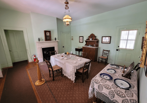 A vintage dining room with a lace tablecloth, wooden chairs, a fireplace, and a side table set for a meal.