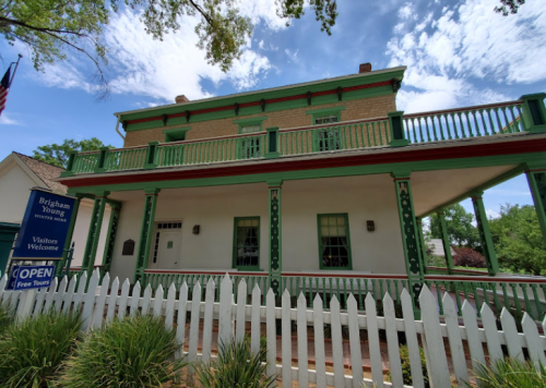 Historic Brigham Young home with green trim, white fence, and open sign, surrounded by trees and blue sky.