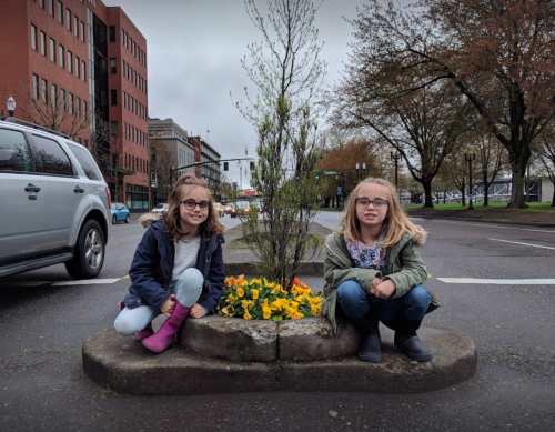 Two girls sit on a concrete planter with flowers, surrounded by a city street and buildings on a cloudy day.