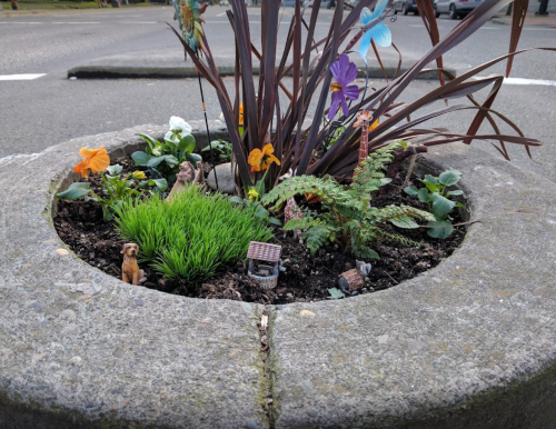 A small garden planter with colorful flowers, greenery, and miniature decorations, including a dog and a tiny house.
