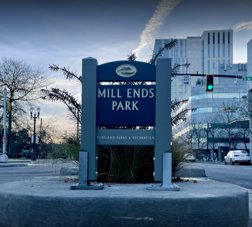 Sign for Mill Ends Park in Portland, surrounded by grass and city buildings, with traffic lights in the background.