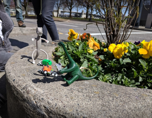 A green dinosaur and a small vehicle toy beside a figure, all placed near colorful flowers in a concrete planter.