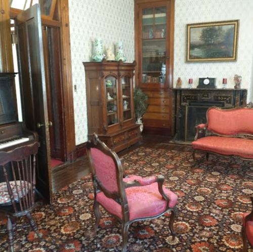 A vintage living room with ornate furniture, a piano, and a decorative fireplace, featuring patterned wallpaper and a rug.