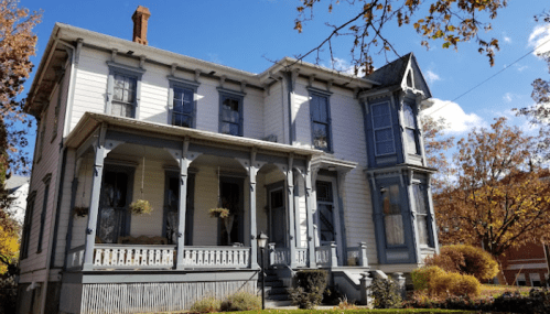 A large, historic two-story house with a gray and white exterior, featuring a wraparound porch and decorative details.