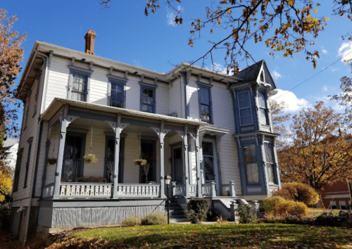 A charming two-story Victorian house with a wraparound porch, surrounded by autumn foliage and a clear blue sky.