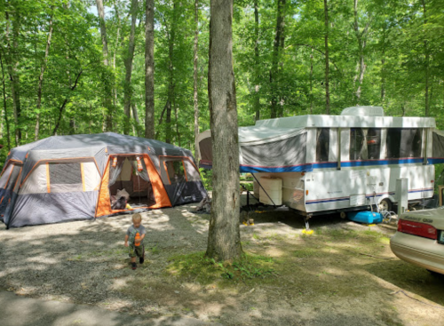 A child walks near a large tent and a camper trailer in a green, wooded campsite.