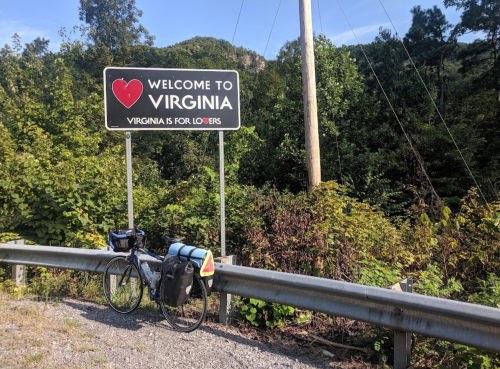 A bicycle with panniers parked beside a "Welcome to Virginia" sign surrounded by greenery.