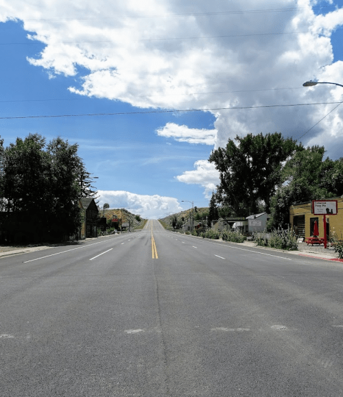 A wide, empty road stretches into the distance under a blue sky with fluffy clouds, flanked by trees and buildings.