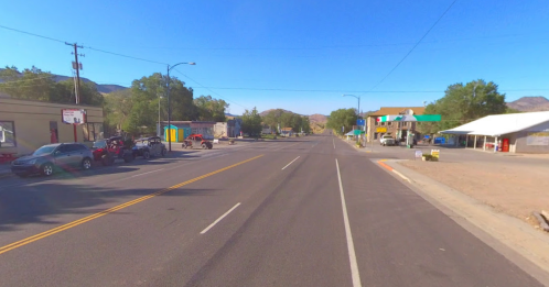 A quiet street scene with shops, parked cars, and mountains in the background under a clear blue sky.