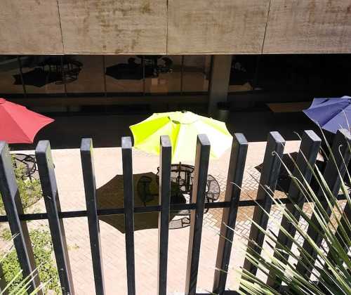 A view from above showing colorful umbrellas over outdoor tables in a courtyard, with a fence in the foreground.