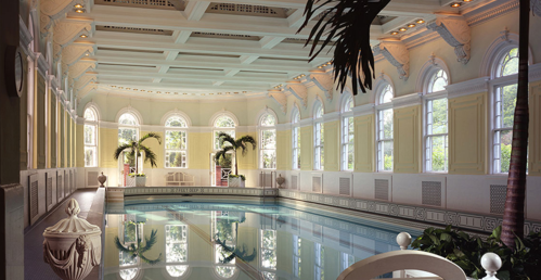 Elegant indoor pool surrounded by large windows and palm trees, featuring ornate architectural details and soft lighting.